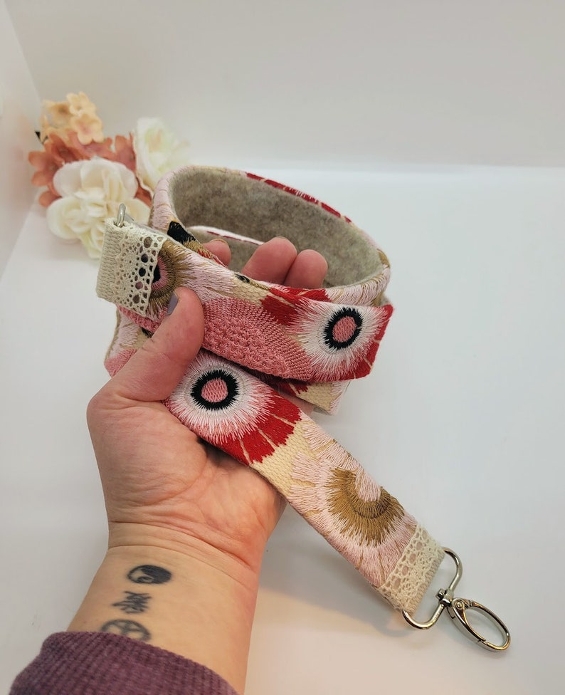 May include: A pink and red floral patterned fabric strap with a white lace trim and a silver metal key ring. The strap is folded in half and held in a hand.