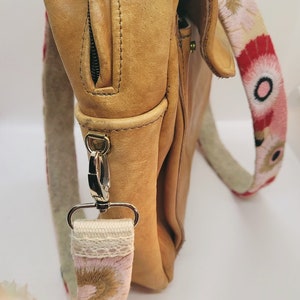 May include: A tan leather satchel bag with a floral patterned strap. The strap is attached to the bag with a silver clasp.