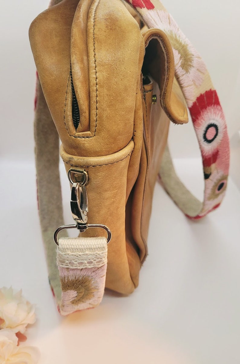 May include: A tan leather satchel bag with a floral patterned strap. The strap is attached to the bag with a silver clasp.