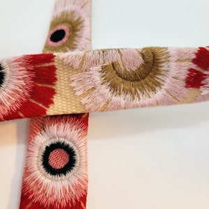 May include: Close-up of a beige fabric with a floral pattern. The fabric has red, pink, and brown flowers with white centers. The flowers are embroidered with a fluffy texture.