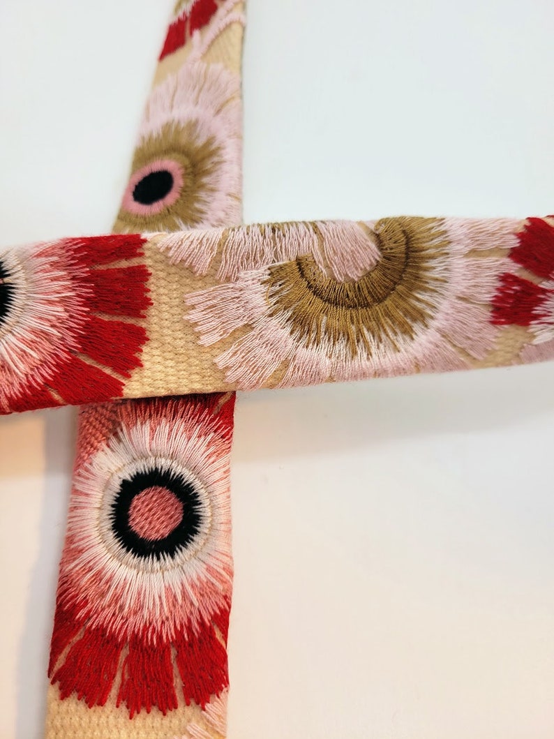 May include: Close-up of a beige fabric with a floral pattern. The fabric has red, pink, and brown flowers with white centers. The flowers are embroidered with a fluffy texture.