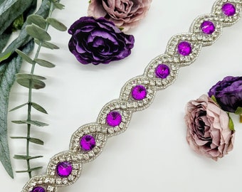 May include: A silver and purple beaded trim with a wavy design. The trim is decorated with small, round, purple rhinestones.