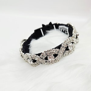 May include: A black pet collar with a sparkling, ornate design. The collar is encrusted with numerous clear rhinestones, creating a dazzling effect. The collar is set against a white, fluffy background.