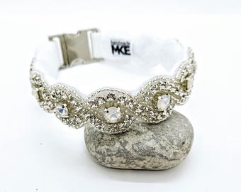 Könnte beinhalten: Weißes Stoffarmband mit einer silbernen Schnalle und einer Reihe von klaren Strasssteinen. Das Armband hat ein wellenförmiges Design und der Text "remarkable MKE" ist auf den Stoff gedruckt.