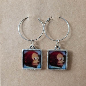May include: A pair of silver hoop earrings with square pendants. Each pendant features a cartoon image of a monkey's face with red hair and a light skin tone, set against a blue background.