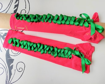 Puede incluir: Un par de calentadores de brazo de color rosa con cinta de satén verde atada en lazos en los extremos. La cinta está tejida en la tela de los calentadores de brazo.