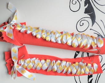 Puede incluir: Dos calienta brazos de tela roja con una cinta blanca atada alrededor. La cinta está decorada con un patrón de patitos de goma amarillos.