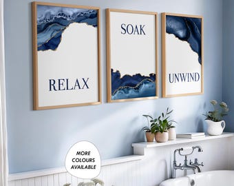 May include: Three framed prints with blue and gold abstract designs. The prints have the words "Relax", "Soak", and "Unwind" on them. The prints are hanging on a light blue wall above a white bathtub.