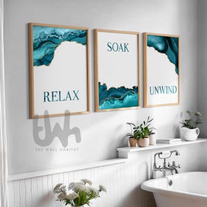 May include: Three framed prints with blue and gold abstract designs. The prints have the words "Relax", "Soak", and "Unwind" on them. The prints are hanging on a white wall above a white bathtub.