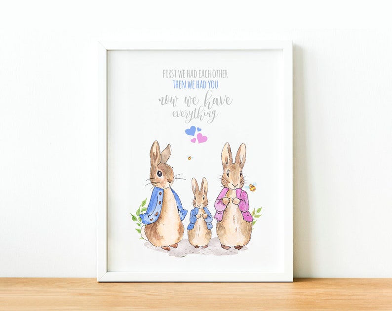 May include: A white framed print with a watercolor illustration of three bunnies. The bunnies are wearing clothes and there are small pink and blue hearts above them. The text reads "First we had each other, then we had you, now we have everything."