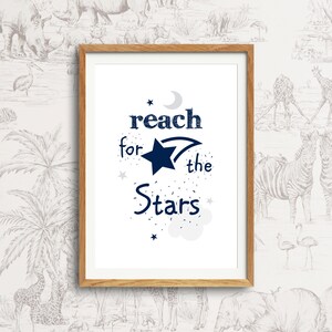 以下が含まれることがあります： 白い背景にネイビーブルーの文字で「reach for the stars」と書かれた額装されたプリント。テキストには三日月、流れ星、小さな星がいくつか添えられています。
