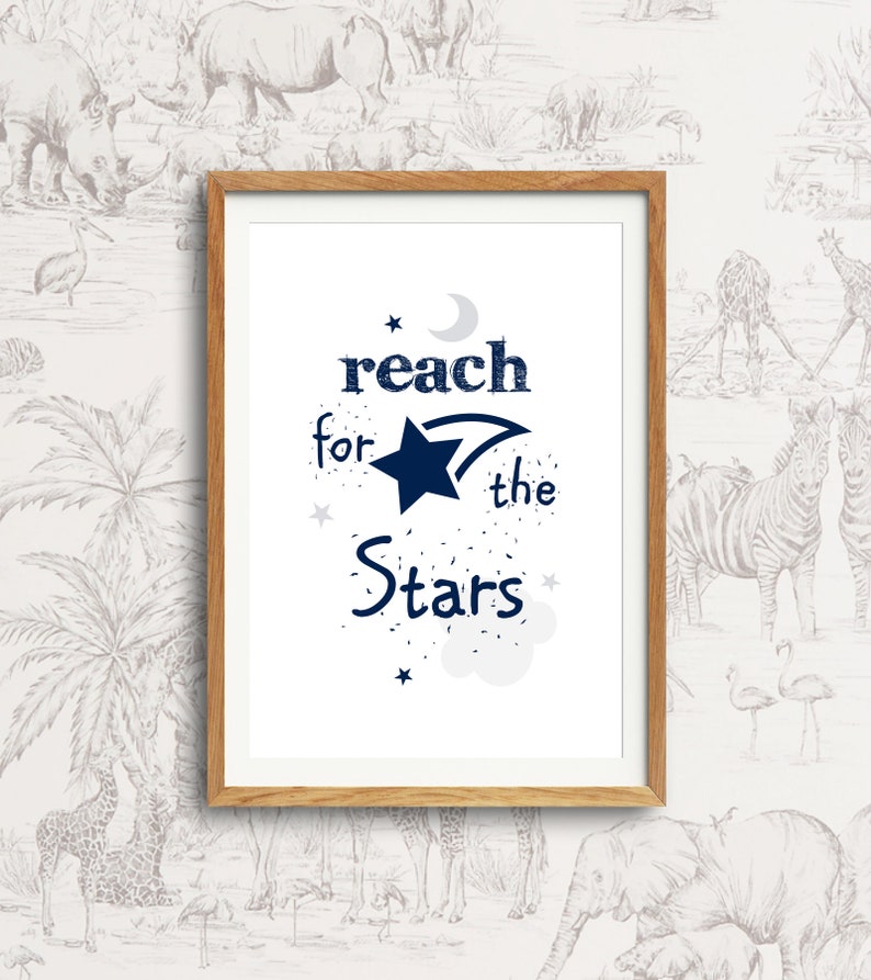 以下が含まれることがあります： 白い背景にネイビーブルーの文字で「reach for the stars」と書かれた額装されたプリント。テキストには三日月、流れ星、小さな星がいくつか添えられています。