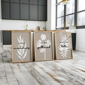 Op de afbeelding: Drie ingelijste kunstprints met botanische ontwerpen en tekst. De prints hebben een neutraal kleurenpalet met witte bladillustraties en zinnen als "THE BEST memories", "ARE MADE gathered" en "AROUND THIS table". De frames zijn licht van kleur.