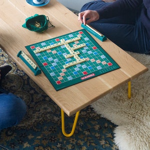 May include: Two people playing a game of Scrabble on a wooden coffee table with yellow hairpin legs. The game board is green with a blue and white grid. The tiles are white with black letters. The players are sitting on a patterned rug.