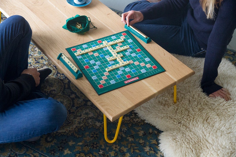 May include: Two people playing a game of Scrabble on a wooden coffee table with yellow hairpin legs. The game board is green with a blue and white grid. The tiles are white with black letters. The players are sitting on a patterned rug.