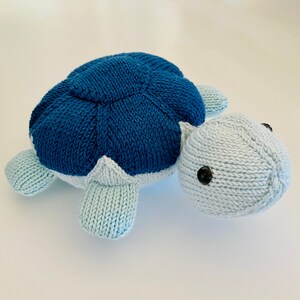 Peut inclure: Un jouet en forme de tortue en tricot bleu et bleu clair. La tortue a une carapace détaillée et des yeux noirs.