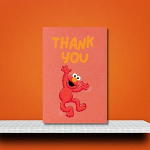 K&ouml;nnte beinhalten: Eine korallenfarbene Gru&szlig;karte mit den Worten &bdquo;THANK YOU&ldquo; in Gelb. Die Karte zeigt eine rote Cartoonfigur mit wei&szlig;en Augen und einem l&auml;chelnden Gesichtsausdruck in einer Tanzpose. Die Karte befindet sich auf einem wei&szlig;en Regal vor einem orangefarbenen Hintergrund.