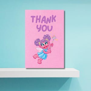 K&ouml;nnte beinhalten: Rosa Karte mit den Worten "THANK YOU" in Lila. Die Karte zeigt eine Cartoon-Fee mit lila Haaren, einem blauen Kleid und Fl&uuml;geln, die einen Zauberstab mit Sternen h&auml;lt. Die Karte befindet sich auf einem wei&szlig;en Regal vor einer hellblauen Wand.