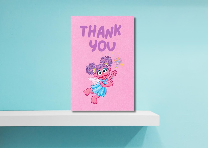 K&ouml;nnte beinhalten: Rosa Karte mit den Worten "THANK YOU" in Lila. Die Karte zeigt eine Cartoon-Fee mit lila Haaren, einem blauen Kleid und Fl&uuml;geln, die einen Zauberstab mit Sternen h&auml;lt. Die Karte befindet sich auf einem wei&szlig;en Regal vor einer hellblauen Wand.