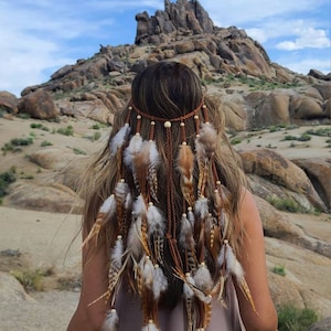 Pode incluir: Uma mulher com cabelo castanho comprido usando uma faixa de cabeça de couro marrom e penas com penas brancas e marrons. A faixa de cabeça é adornada com pequenas contas de madeira. A mulher está em pé em uma paisagem desértica com colinas rochosas ao fundo.
