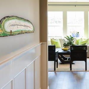 May include: A wall-mounted key holder made from a slice of agate stone. The key holder features a natural green, white, and brown color palette with brass hooks. The agate slice is mounted on a wall with a white wainscoting.