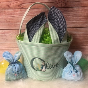 May include: A green and white striped Easter basket with bunny ears, personalized with the name "Olive". The basket is filled with green grass and includes two blue bunny-shaped bags.