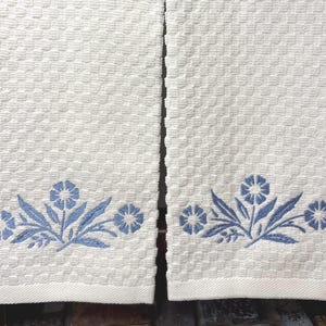 May include: Two white kitchen towels with a blue embroidered floral design. The design features stylized flowers with leaves.