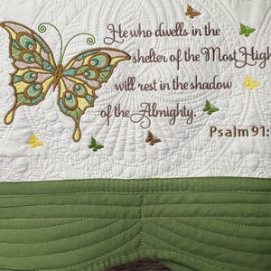 May include: A white quilted textile featuring a large butterfly with green, yellow, and brown details. The text "He who dwells in the shelter of the Most High will rest in the shadow of the Almighty. Psalm 91:1" is embroidered in brown.