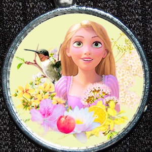 Puede incluir: Un colgante redondo de plata con una imagen colorida de una princesa de dibujos animados con el pelo largo y rubio, rodeada de flores y un colibrí. El fondo es de color amarillo claro.