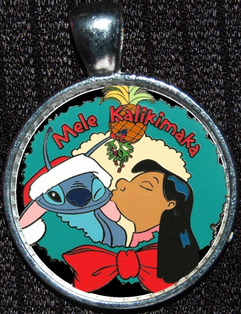 Puede incluir: Un amuleto de plata con una imagen colorida de Stitch y Lilo de la pel&iacute;cula de Disney Lilo & Stitch. La imagen muestra a Stitch con un gorro de Pap&aacute; Noel y Lilo besando a Stitch en la mejilla. El amuleto tiene el texto "Mele Kalikimaka" alrededor del borde.