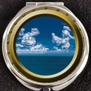 May include: A round, silver compact mirror with a gold-colored frame. The mirror shows a view of the ocean and sky through a porthole. The sky is blue with white clouds that form the shape of two smiling faces.