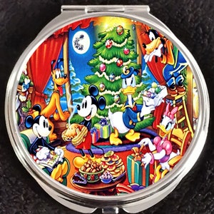 Op de afbeelding: Een ronde compact spiegel met een kleurrijk beeld van Mickey Mouse, Minnie Mouse, Donald Duck, Goofy en Pluto die kerst vieren. De personages zijn verzameld rond een gedecoreerde kerstboom met cadeaus en koekjes.