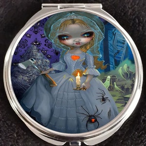 May include: A compact mirror with a gothic-inspired design featuring a pale-skinned doll with blue eyes and blonde hair wearing a white dress and holding a candle and an axe. The background is a dark purple with a spooky mansion and ghosts.