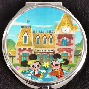 Puede incluir: Un espejo compacto con una ilustración colorida de Mickey y Minnie Mouse haciendo un picnic frente a la estación de tren de Disneyland. El fondo es un cielo azul brillante con nubes blancas.