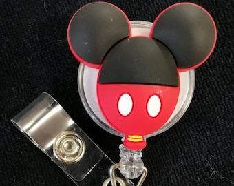 May include: A red and black Mickey Mouse character badge holder with a clear plastic retractable reel. The badge holder has a clear plastic backing and a silver clip.