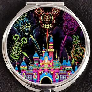Op de afbeelding: Een ronde, zilveren compacte spiegel met een kleurrijk ontwerp. Het ontwerp bevat een levendig kasteel, vuurwerk en gestileerde gezichten van personages tegen een zwarte achtergrond. Het kunstwerk is in heldere neonkleuren.
