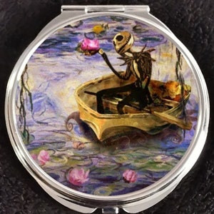 May include: A round, silver compact mirror with a colorful image of a skeleton character in a boat on a water lily pond. The character is holding a pink flower.