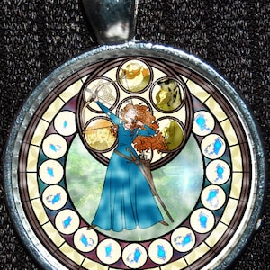 May include: A round, silver-toned pendant with a stained glass design featuring a red-haired woman holding a bow and arrow. The stained glass design also includes a wolf, a bear, and other figures. The pendant is surrounded by a silver-toned frame.
