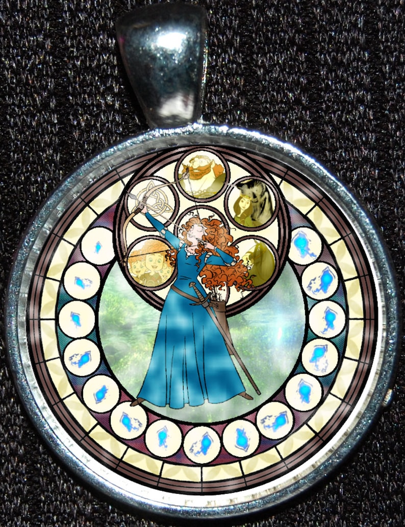 May include: A round, silver-toned pendant with a stained glass design featuring a red-haired woman holding a bow and arrow. The stained glass design also includes a wolf, a bear, and other figures. The pendant is surrounded by a silver-toned frame.