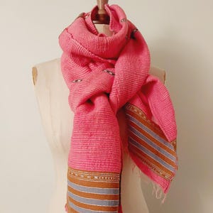 May include: A pink knitted scarf with a textured surface and a gray and gold striped border. The scarf is draped over a dress form.