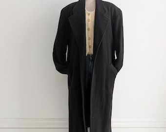 May include: A long black wool coat with a collar and pockets. The coat is on a mannequin and is hanging on a stand.