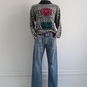 May include: A black and white patterned sweater with a mock neck and three colourful geometric designs. The sweater is worn over a pair of blue jeans.