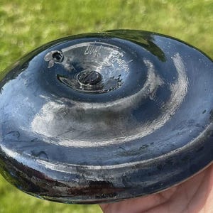 May include: A dark, glossy, oval-shaped glass object with a central screw and a small hole. The surface reflects light, and there is handwritten text on the surface. The object is held in a hand.