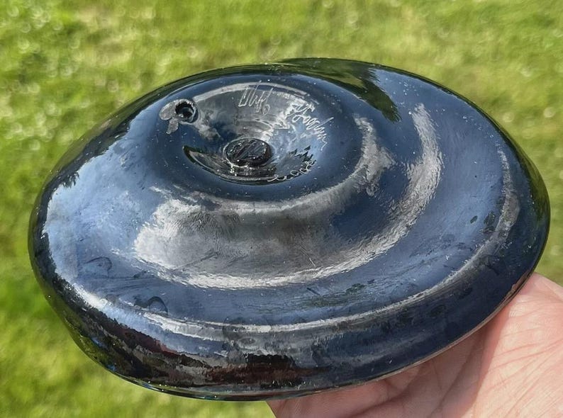 May include: A dark, glossy, oval-shaped glass object with a central screw and a small hole. The surface reflects light, and there is handwritten text on the surface. The object is held in a hand.