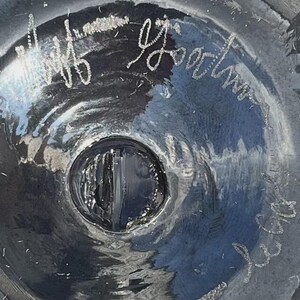 May include: Close-up of a dark, possibly glass, object with a central circular indentation. The surface reflects light, creating highlights and shadows. White script is visible on the surface, including the name "Jeff Goodman".