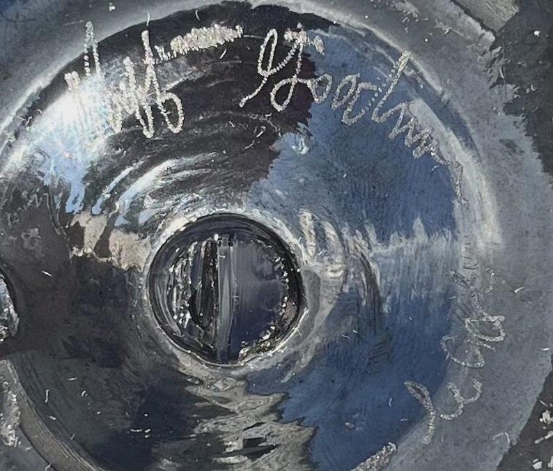 May include: Close-up of a dark, possibly glass, object with a central circular indentation. The surface reflects light, creating highlights and shadows. White script is visible on the surface, including the name "Jeff Goodman".