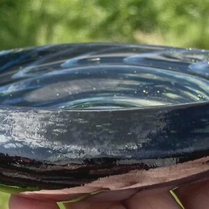May include: A handmade, shallow glass bowl with a dark blue and teal swirl pattern. The bowl has a glossy finish and a slightly irregular shape. The rim is a darker shade of blue. The bowl is held in a hand.