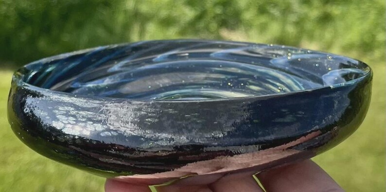 May include: A handmade, shallow glass bowl with a dark blue and teal swirl pattern. The bowl has a glossy finish and a slightly irregular shape. The rim is a darker shade of blue. The bowl is held in a hand.