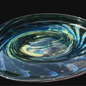 May include: A clear glass decorative bowl with a galaxy design. The bowl features swirling patterns of blue, green, and yellow, resembling a nebula or a spiral galaxy. The bowl is oval-shaped.