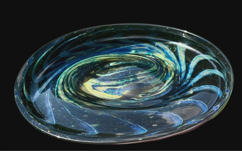 May include: A clear glass decorative bowl with a galaxy design. The bowl features swirling patterns of blue, green, and yellow, resembling a nebula or a spiral galaxy. The bowl is oval-shaped.